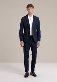 Costume marine en tissu texturé, veste ajustée avec revers en pointe, assortie à une chemise blanche et des chaussures sombres.