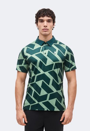 Mann trägt ein kurzärmeliges grünes Polohemd mit dunkelgrünen geometrischen eckigen Mustern, steht vor einem einfarbigen Hintergrund.