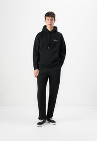 Young man wearing a black hoodie, black pants, and black sneakers, standing against a plain white background with one hand in pocket.
