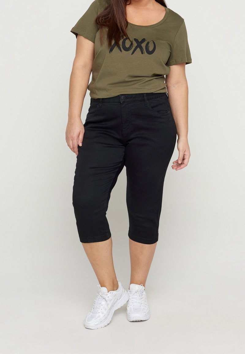 Oliivivihreä T-paita, jossa on "XOXO" -printti, yhdistettynä mustiin capri-housuihin. Housuissa on vartalonmyötäinen malli ja sileä kangas, ja asusteena valkoiset lenkkarit.