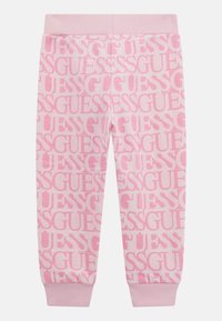 Un pantalon de survêtement rose avec un logo "GUESS" en rose clair qui se répète. Il présente une taille et des poignets côtelés pour un ajustement confortable. Texture de tissu douce.