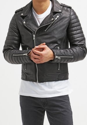 Mann trägt schwarze gesteppte Lederjacke mit Reißverschlussdetails über weißem Hemd und dunkler Hose, Hände in der Taille verschränkt.