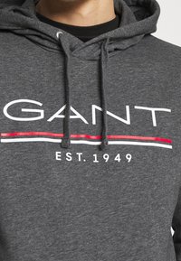 Felpa con cappuccio grigio scuro con logo GANT, strisce rosse e bianche e testo "EST. 1949" sul petto.