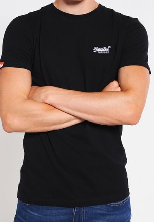 Personne aux bras croisés portant un t-shirt noir SuperDry et un jean bleu sur un fond uni.