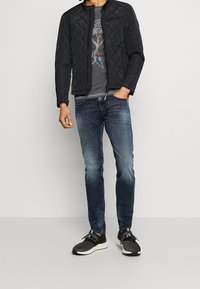 Gepatchte schwarze Jacke mit Stehkragen, grauem Grafik-T-Shirt darunter, blauen Slim-Fit-Jeans und schwarzen Sportschuhen mit weißen Sohlen.