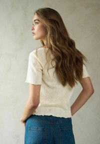 Kortärmad, krämfärgad topp med strukturerade ränder och vågig fåll, i kombination med högmidjade blå denimjeans. Bakvy.