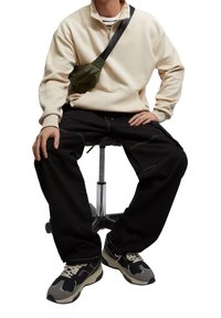 Beige sweatshirt med halv lynlås, sorte løse bukser med hvide syninger, grøn hoftepose og grå-sorte sneakers med lilla detaljer, siddende på en skammel.