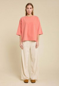 Haut en coton oversized corail avec un col rond, associé à un pantalon large beige et des mules marron, sur un fond neutre.