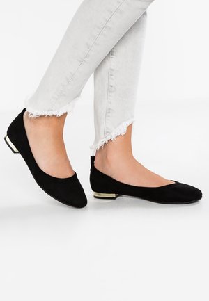 Black suede ballet flats with a rounded toe and a small metallic accent on the heel, worn with light gray frayed jeans.