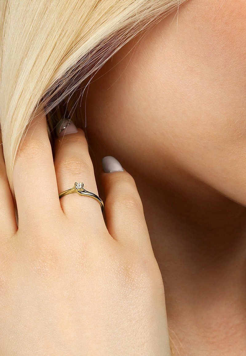 Goldring mit einem gedrehten Design, das einen einzelnen klaren Stein enthält. Wird an einer Hand mit heller Haut und kurzen Nägeln getragen, sanfte Textur.