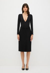 Musta pitkähiha bodysuit syvällä V-päällä, yhdistettynä polven mittaiseen mustaan hameeseen, jossa on vyöllinen vyötärö ja etuosassa laskostus.