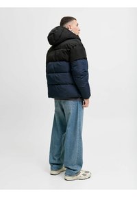 Polstret jakke med en sort øverste del og marineblå nederste del, med hætte, lynlås og struktureret overflade, båret over løse blå jeans.