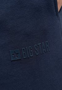 Jūras spēku zila tekstila auduma ar mīkstu tekstūru, kas iezīmēta ar paaugstinātu tumši zilu logu ar tekstu "BIG STAR" un zvaigznes simbolu.