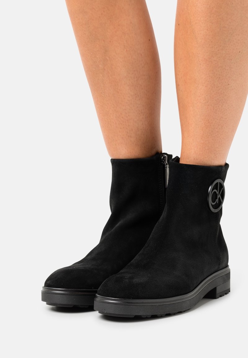 Stivali alla caviglia in suede nero con accento del logo CK rotondo, cerniera laterale, suola piatta e tomaia texturizzata, che offrono un design elegante e moderno.