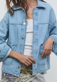 Femme portant une veste en denim bleu clair par-dessus un débardeur blanc et une jupe à motifs, ajustant la veste avec les mains près des poches.
