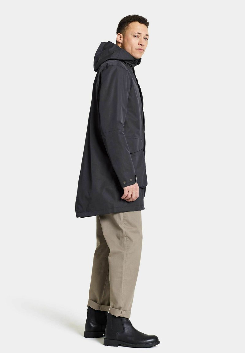 Langer schwarzer wasserdichter Mantel mit Kapuze, der eine gerade Passform hat, Druckknöpfe zum Schließen und seitliche Taschen bietet. Kombiniert mit beigen Hosen und schwarzen Stiefeln.