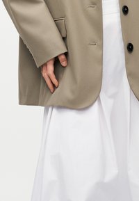 Το χέρι ακουμπά κοντά στην τσέπη ενός ταμπά σακακιού που φοριέται πάνω από μια λευκή ρέουσα φούστα ή φόρεμα, εστιάζοντας στις λεπτομέρειες του μανικιού και του κάτω κορμού.