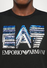 Tuvumā melns krekls ar ziliem, sudraba un baltiem pērļu virpinājumiem ar uzrakstu "EA7" un baltu "EMPORIO ARMANI" tekstu ar logotipu zem kakla apgriezuma.