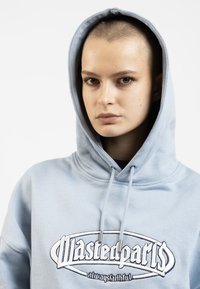 Šviesiai mėlynas džemperis su raišteliu ir dideliu logotipu, pagamintas iš minkštos medžiagos. Turi erdvią kapučą ir laisvo kirpimo dizainą.