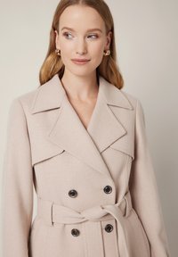 Jasno taupe'owy płaszcz wełniany z podwójnym zapięciem, z kołnierzem z klapami i czarnymi guzikami. Posiada pasek do wiązania w talii dla regulacji.