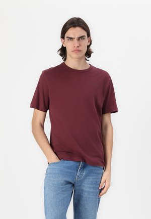 Bordeauxfarbenes T-Shirt mit kurzen Ärmeln, runder Halsauschnitt, weicher Baumwollstoff, präsentiert mit blauen Jeans. Das Model steht vor einem schlichten weißen Hintergrund.