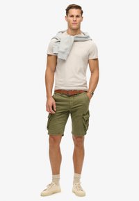 Tan t-shirt, olive green cargo shorts with side pockets, brown belt, gray sweatshirt draped over shoulders, beige sneakers, and patterned socks.