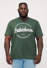 Grünes Baumwoll-T-Shirt mit einem weißen Druck, der die Designs "Jack & Jones" und "Denim Supply Co." zeigt. Kurze Ärmel, lockere Passform und glatte Textur.