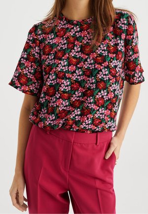 Femme portant un haut noir à manches courtes avec imprimé floral rouge et rose et un pantalon taille haute uni rose foncé, debout avec une main dans la poche.