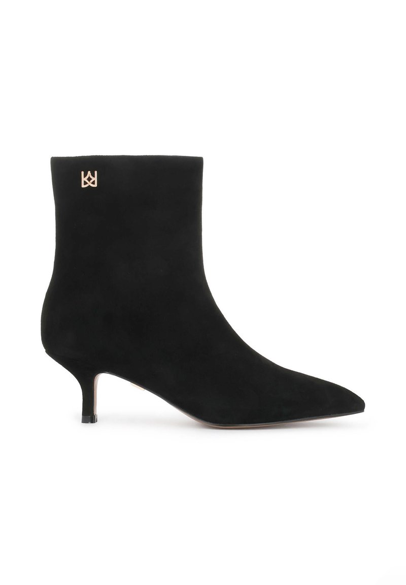 Bota de tobillo de ante negra con puntera afilada y tacón de aguja. Presenta un acento de logo dorado cerca de la parte superior. Textura suave y mate.