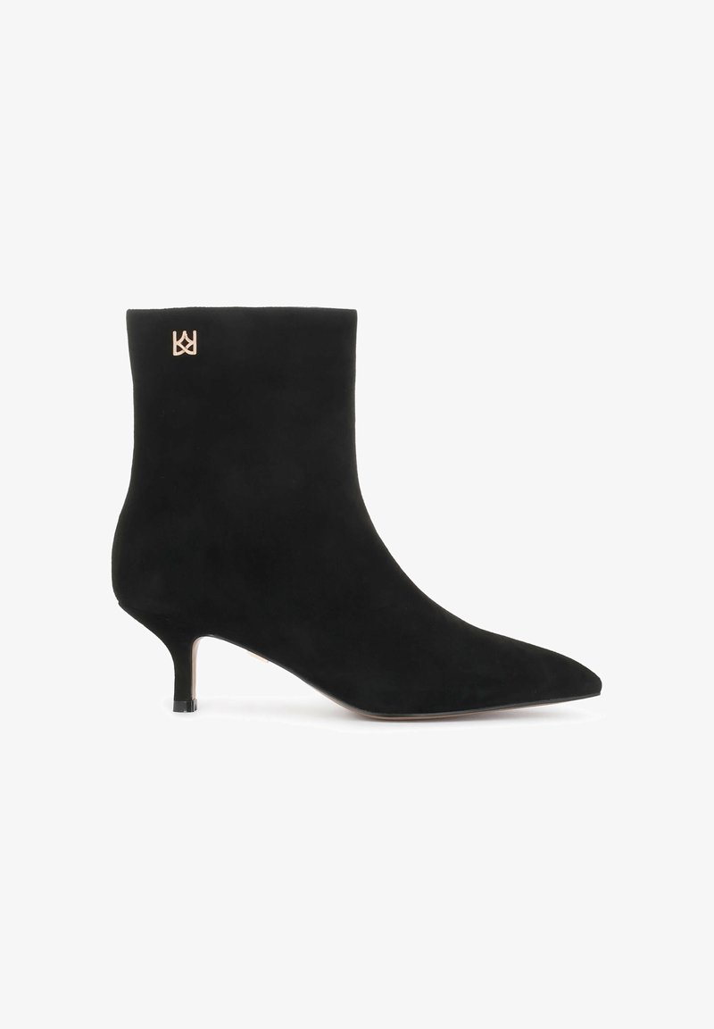 Bota de tobillo de ante negra con puntera afilada y tacón de aguja. Presenta un acento de logo dorado cerca de la parte superior. Textura suave y mate.