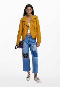 Gul läderbikerjacka med silverdragkedjor, kombinerad med blå patchwork-jeans med olika texturer och en svart väska med en accent.