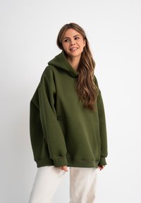 Haut à capuche oversize vert olive avec une grande capuche, une poche frontale et des poignets côtelés, fabriqué en tissu doux. Associé à un pantalon clair.