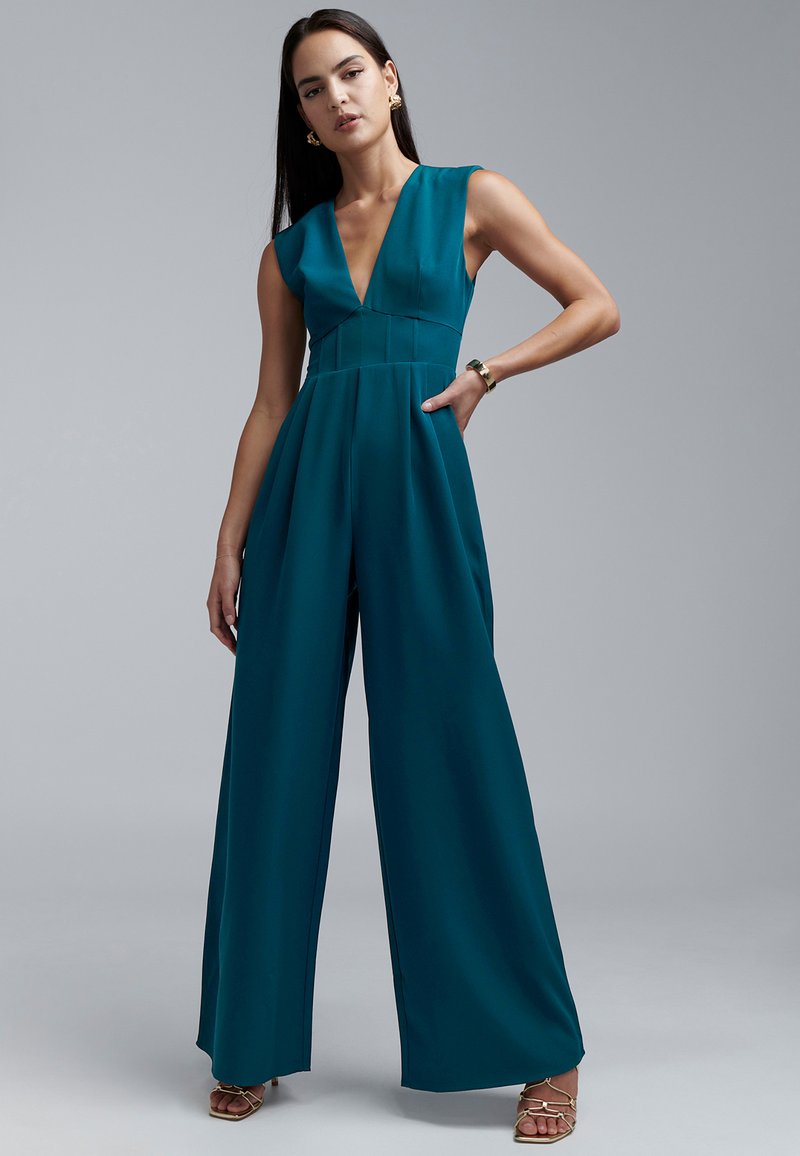 Blauer Overall mit tiefem V-Ausschnitt, ärmellosem Design, weitem Bein und plissiertem Detail. Glatter Stoff und minimalistische Hardware-Akzente.