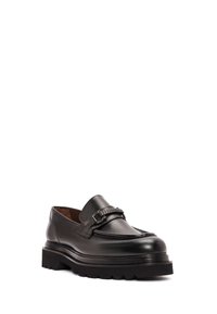 Mocasín de cuero negro con una suela gruesa, que presenta un acento trenzado y un detalle de metal en la parte delantera. Textura suave con un acabado pulido.