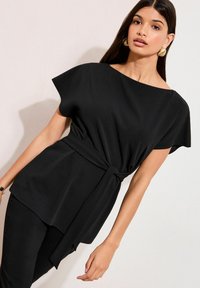 Sort top med korte ærmer, en bådudskæring og et knyttet taljebånd. Fremstillet af glat stof med en flydende silhuet og asymmetrisk kant.