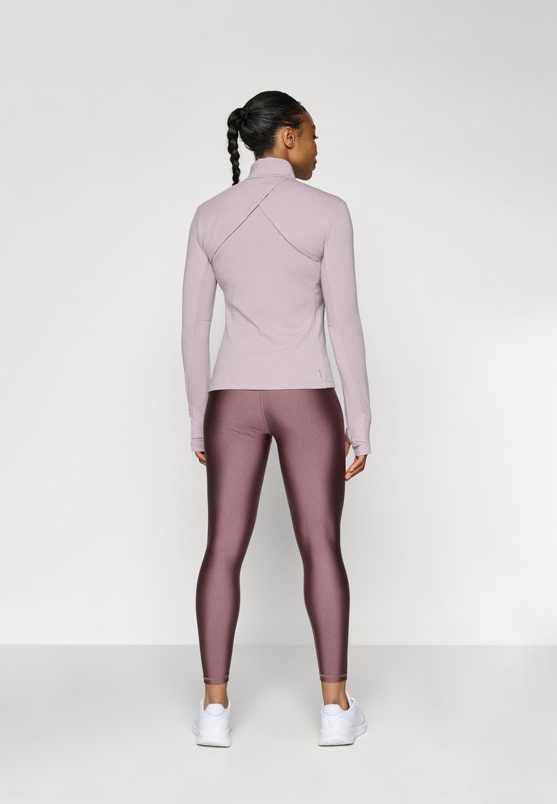 Eine Frau mit geflochtenem Haar steht mit dem Rücken zu uns, trägt ein helllila langärmliges Oberteil, glänzende mauve Leggings und weiße Sportschuhe.