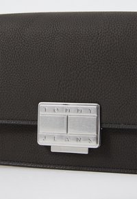 Portafoglio in pelle nera con texture, dotato di una chiusura rettangolare argentata incisa con la scritta "TOMMY JEANS" e un design rettangolare diviso.