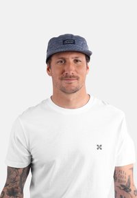 Casquette grise à motifs avec un patch logo à l'avant, portée avec un t-shirt blanc uni. Présente des tatouages visibles sur les bras.