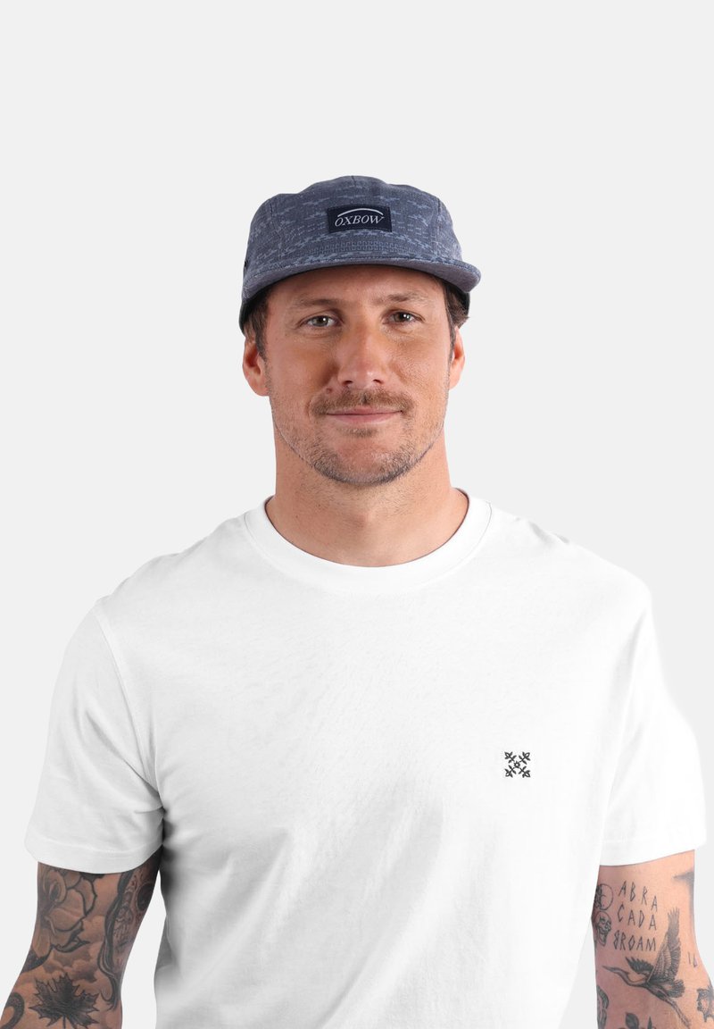Casquette grise à motifs avec un patch logo à l'avant, portée avec un t-shirt blanc uni. Présente des tatouages visibles sur les bras.
