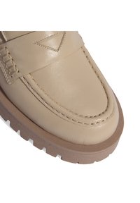 Mocassin en cuir beige avec des coutures visibles et un accent triangulaire. Il dispose d'une épaisse semelle en caoutchouc brun avec une texture antidérapante.