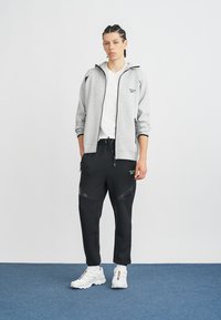 Szürke zipzáras kapucnis póló fekete részletekkel, fekete farmernadrággal, amely texturált panelekből áll. Fehér sportcipővel kiegészítve.