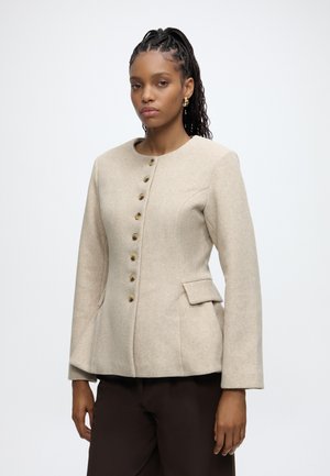 Vrouw met gevlochten haar die een beige button-up jas en donkere broek draagt, staand tegen een effen lichte achtergrond.