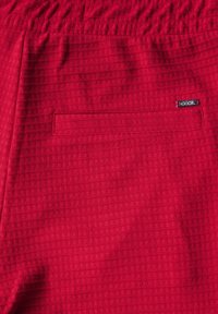 Rood textiel met een geweven patroon, voorzien van een elastische tailleband en een enkele achterzak. Kleine zwarte logo-tag aanwezig.