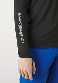 Juodas ilgas rankovės rash guardas su lygiu paviršiumi ir sidabriniu "Quiksilver" logotipu ant rankos, dėvimas su mėlynomis maudymosi šortais.