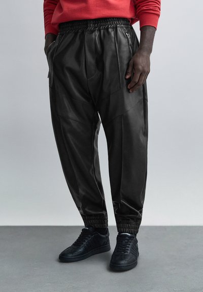 Μαύρο δερμάτινο παντελόνι jogger με ελαστική μέση, τσέπες μπροστά με φερμουάρ και ριμπ μανσέτες στους αστραγάλους. Συνδυασμένο με μαύρα αθλητικά παπούτσια.