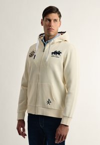 Sudadera crema con cremallera y capucha, presenta logotipos y detalles en azul marino, bolsillos frontales y puños y dobladillo de punto. La tela parece suave y texturizada.