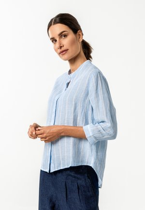 Frau mit zurückgebundenem dunklem Haar trägt eine hellblaue, langärmelige, gestreifte Bluse mit Knöpfen und dunkelblaue Hosen, steht vor einem weißen Hintergrund.