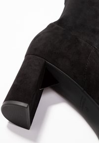 Botte à talons hauts en suède noir avec un accent métallique triangulaire sur le talon, surface structurée et bout arrondi. Semelle en caoutchouc avec des rainures horizontales.