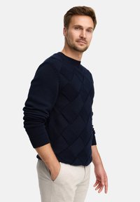 Granatowy sweter z fakturowanym wzorem w romby, okrągłym dekoltem i ściągaczami na rękawach, noszony z jasnoszarymi spodniami.