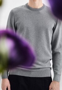 Pull en maille grise avec un col rond, des poignets et un ourlet côtelés. La texture semble douce avec un motif subtil. Les mains du mannequin sont visibles.